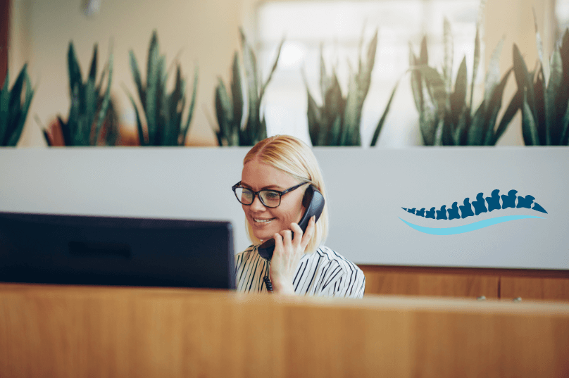 A friendly chiropractic assistant smiling behind a modern clinic reception desk.